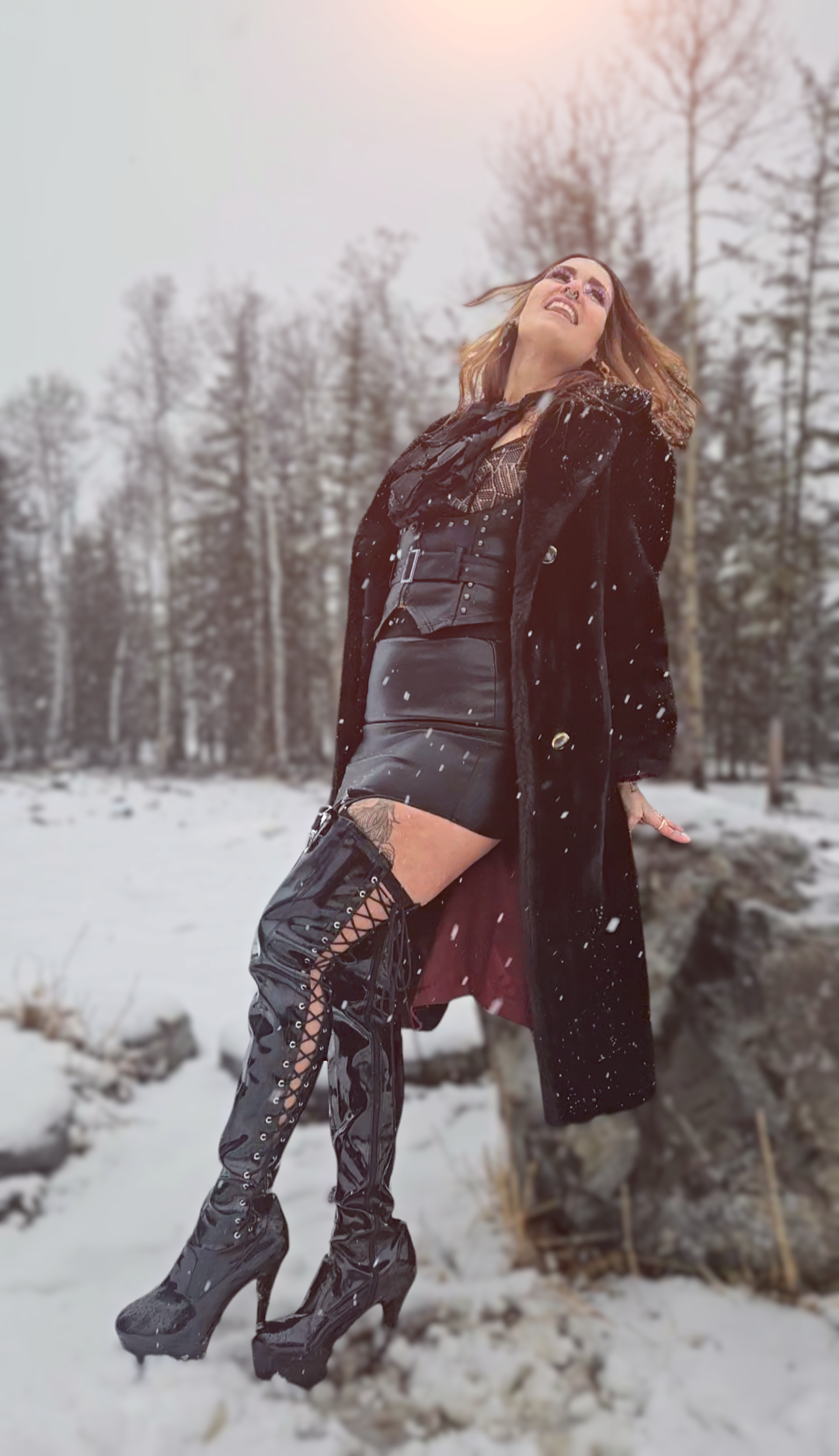 A Domme standing outdoors in a snowy landscape, wearing shiny black thigh-high lace-up boots, a black leather mini skirt, a black corset, and a long black fur coat, with snow falling around her.