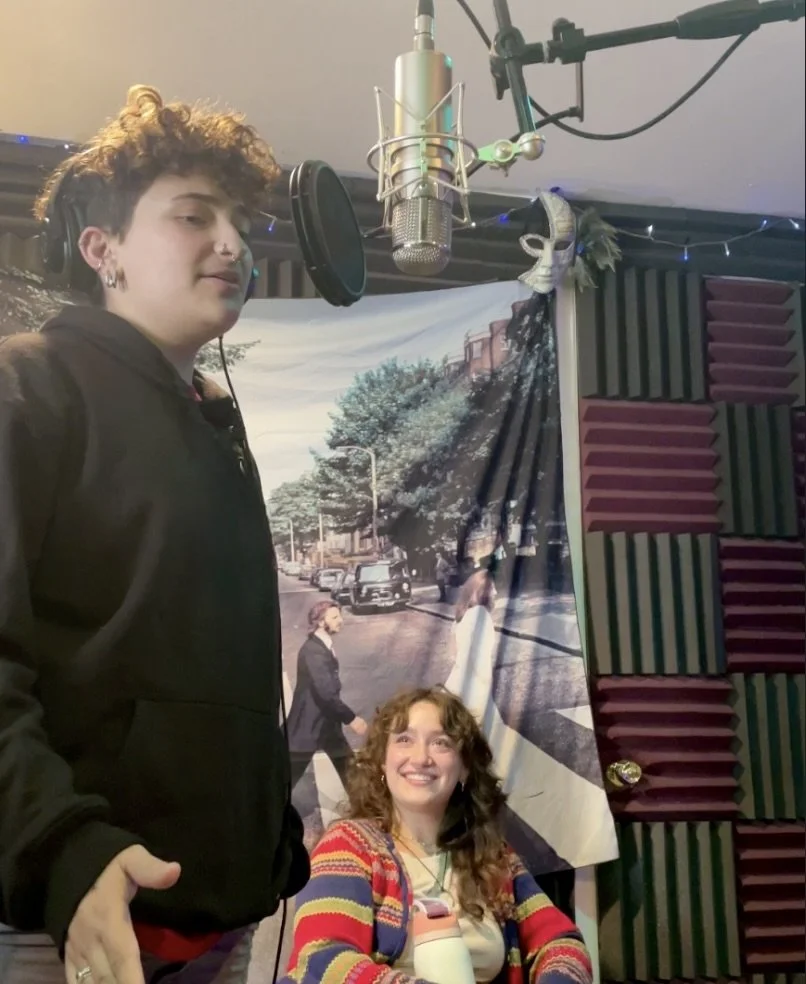 A young man with curly hair and earrings stands in front of a microphone in a recording studio, with a woman sitting and smiling in front of a background depicting The Beatles' Abbey Road album cover.