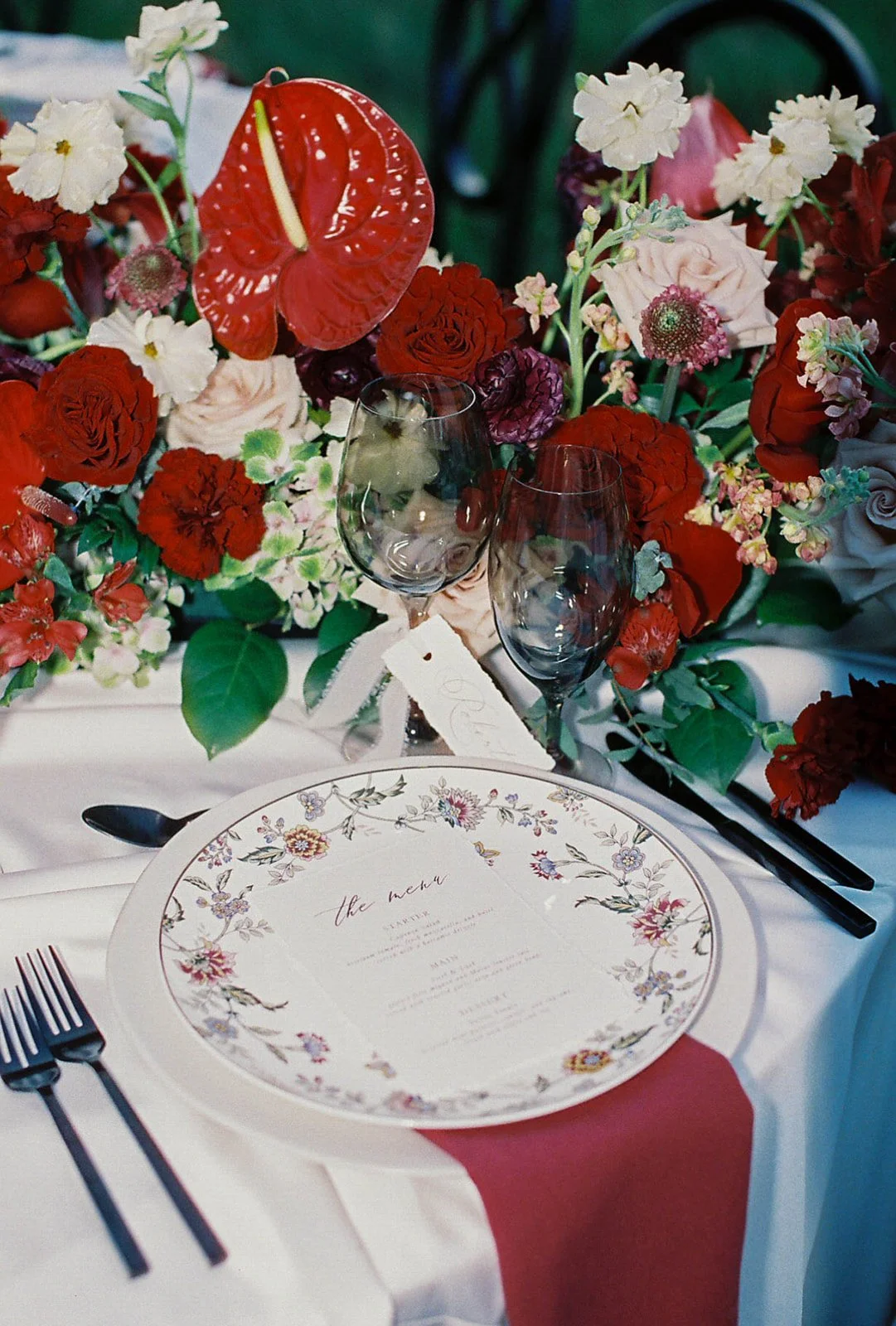 A wedding table setting with floral centerpiece, two smokey wine glasses, a menu, and cutlery, with a red and pink flower arrangement as decoration.
