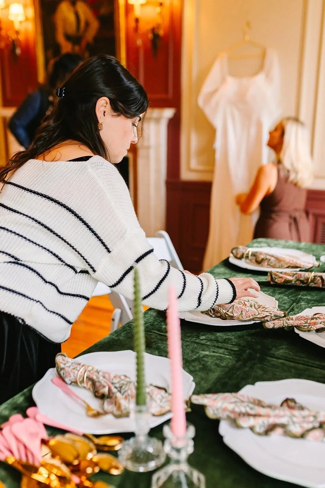 A wedding planner setting a table with plates, napkins, and cutlery, in a decorated indoor space with paintings at Misselwood Events at Endicott College.