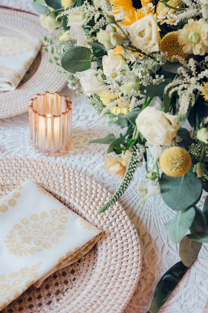 Close-up of a table setting with a woven placemat, floral napkin, pink glass candle holder, and a large bouquet of white, yellow, and green flowers on a white lace tablecloth at the Garden at Elm Bank.