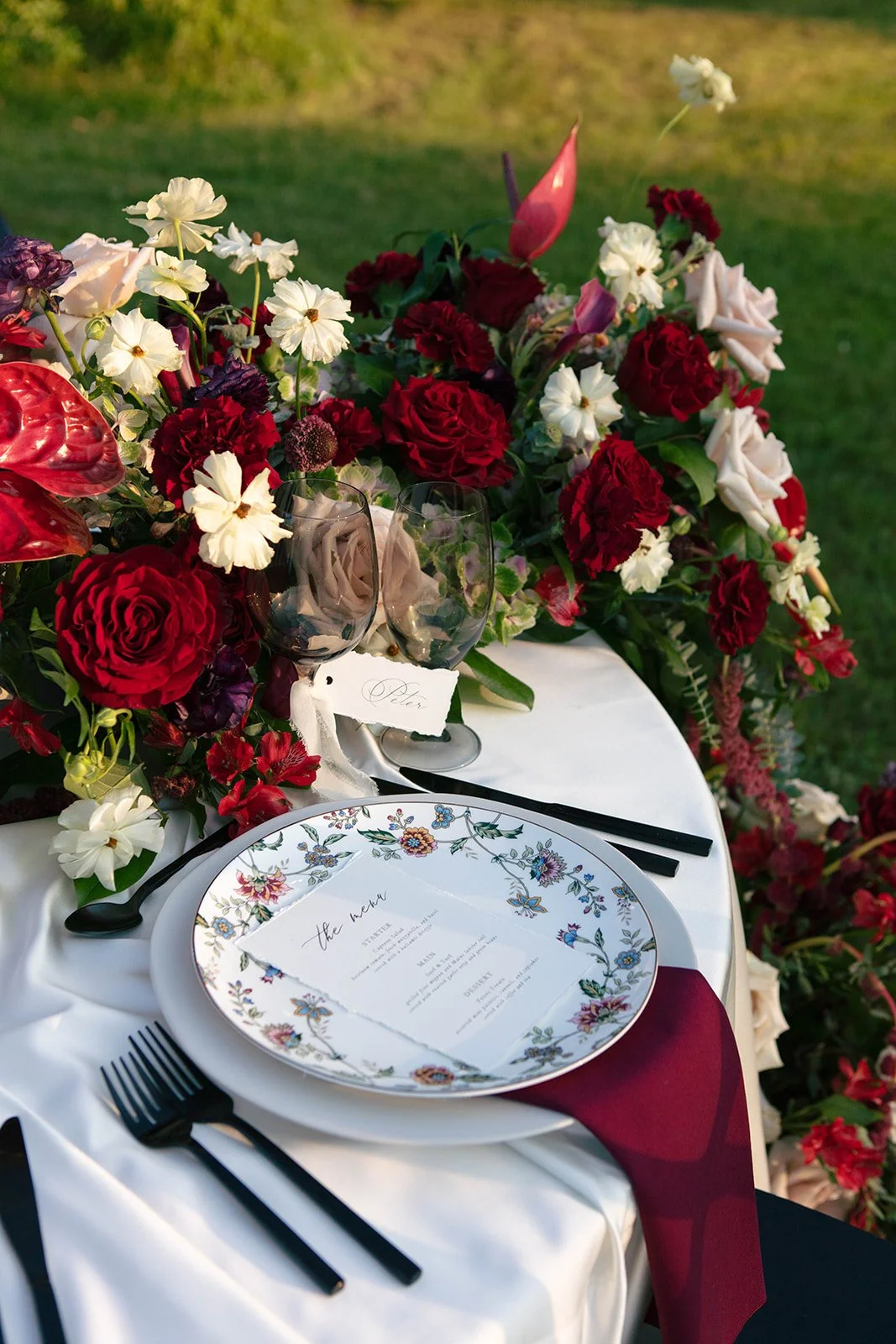 Elegant outdoor dining table decorated with a large floral centerpiece including red, white, and pink flowers, a floral-patterned plate with a menu, black cutlery, and wine glasses, set on a white tablecloth with a red napkin.