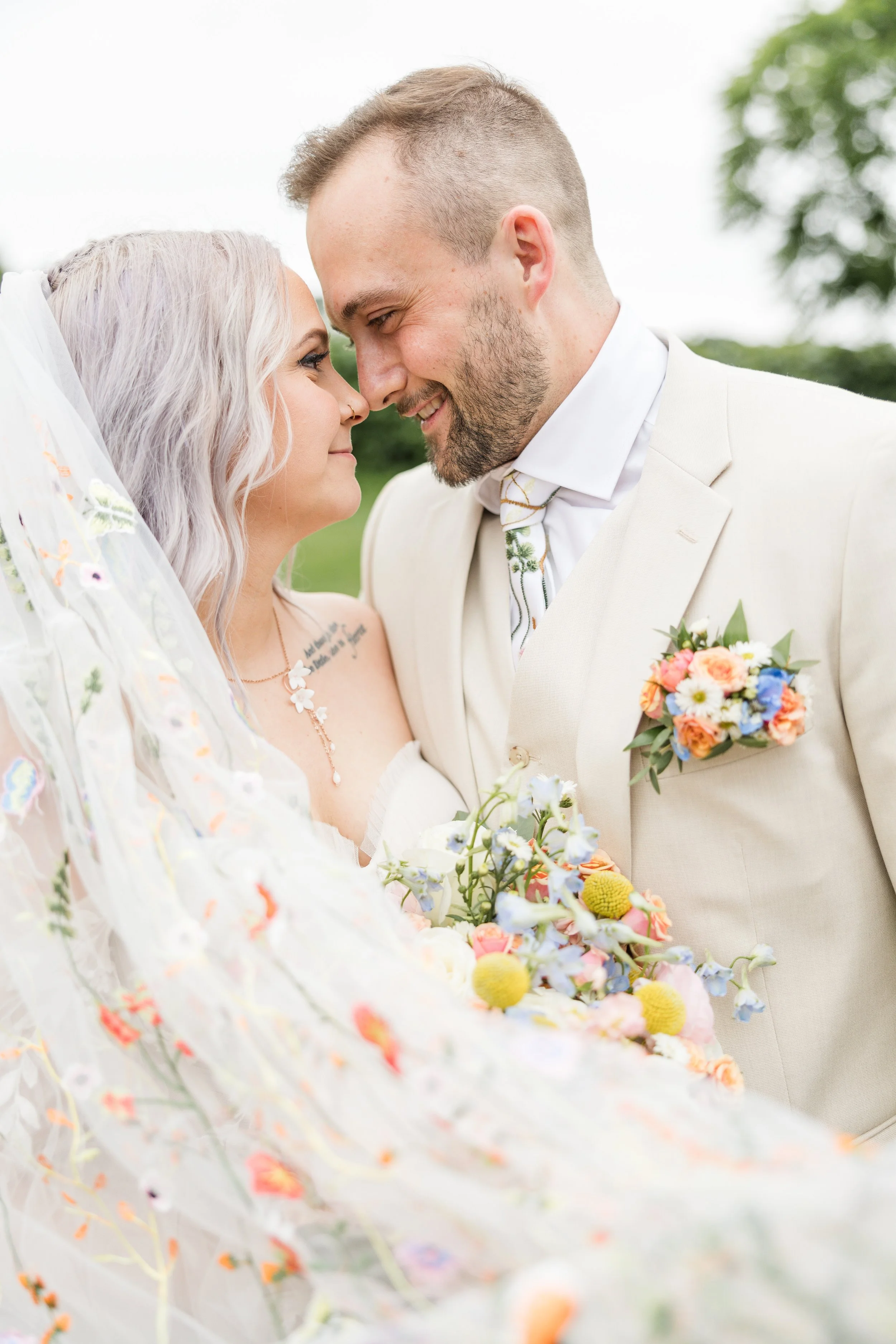 A bride and groom face-to-face, noses touching, smiling outdoors, with the bride holding a bouquet of flowers and a veil with colorful embroidery at Smith Barn in Peabody.