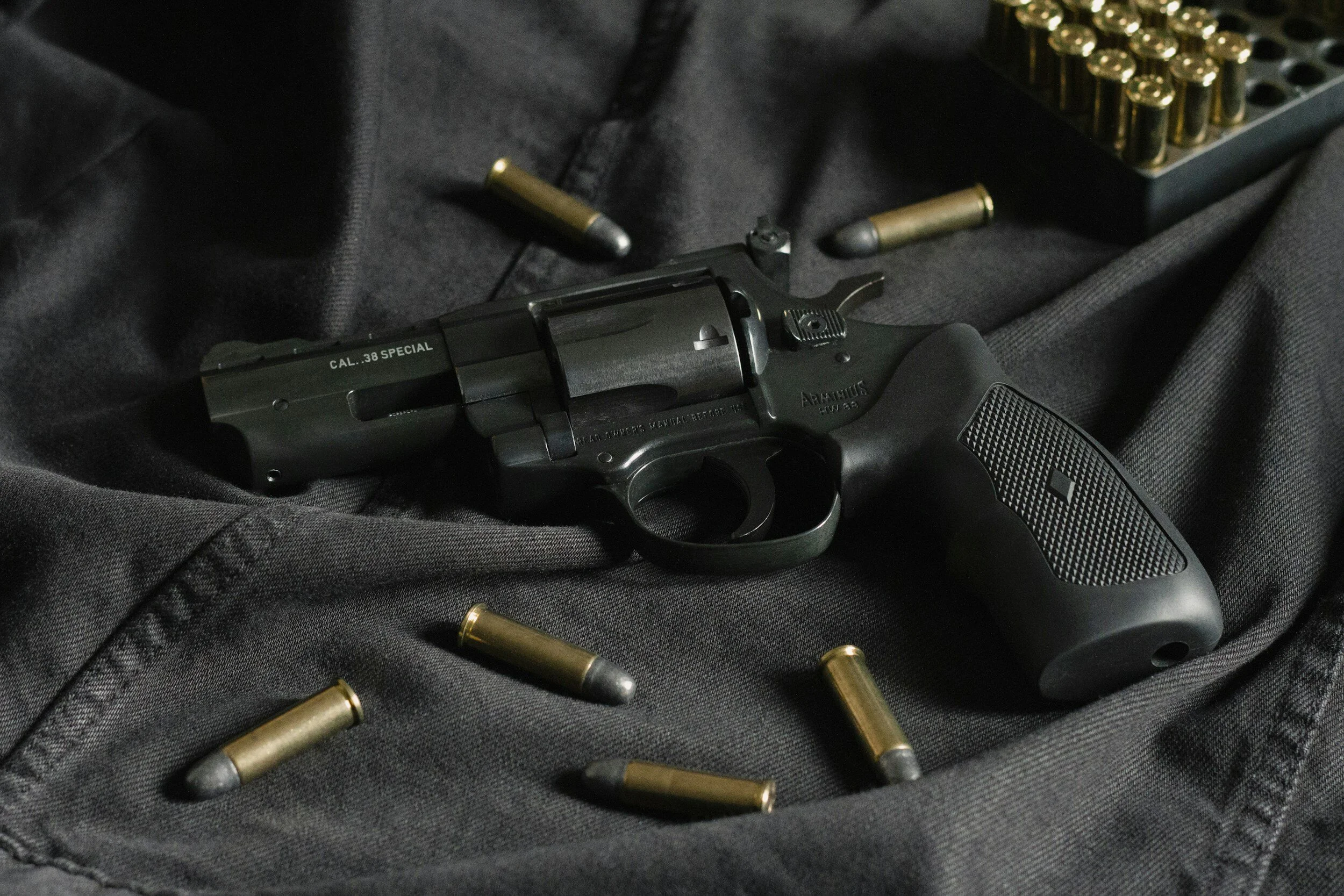 A black revolver with markings CAL.38 SPECIAL, surrounded by several brass and lead bullets, resting on a dark fabric surface.