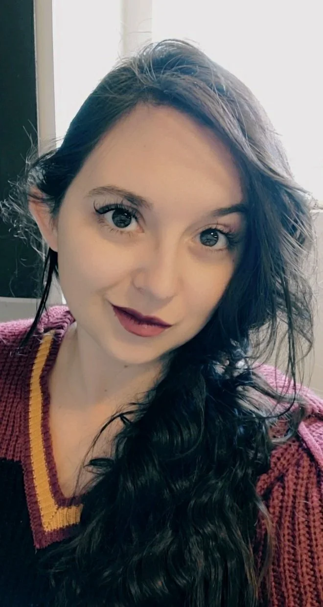 A young woman with long, dark, wavy hair and fair skin, wearing a burgundy sweater with yellow trim, posing indoors with natural light