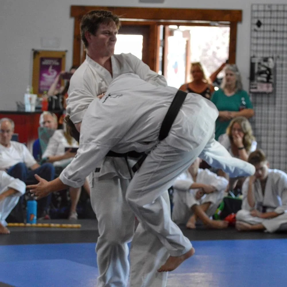Two martial artists wearing white gi uniforms and black belts are practicing judo or Brazilian jiu-jitsu on a blue mat, with spectators watching in the background.