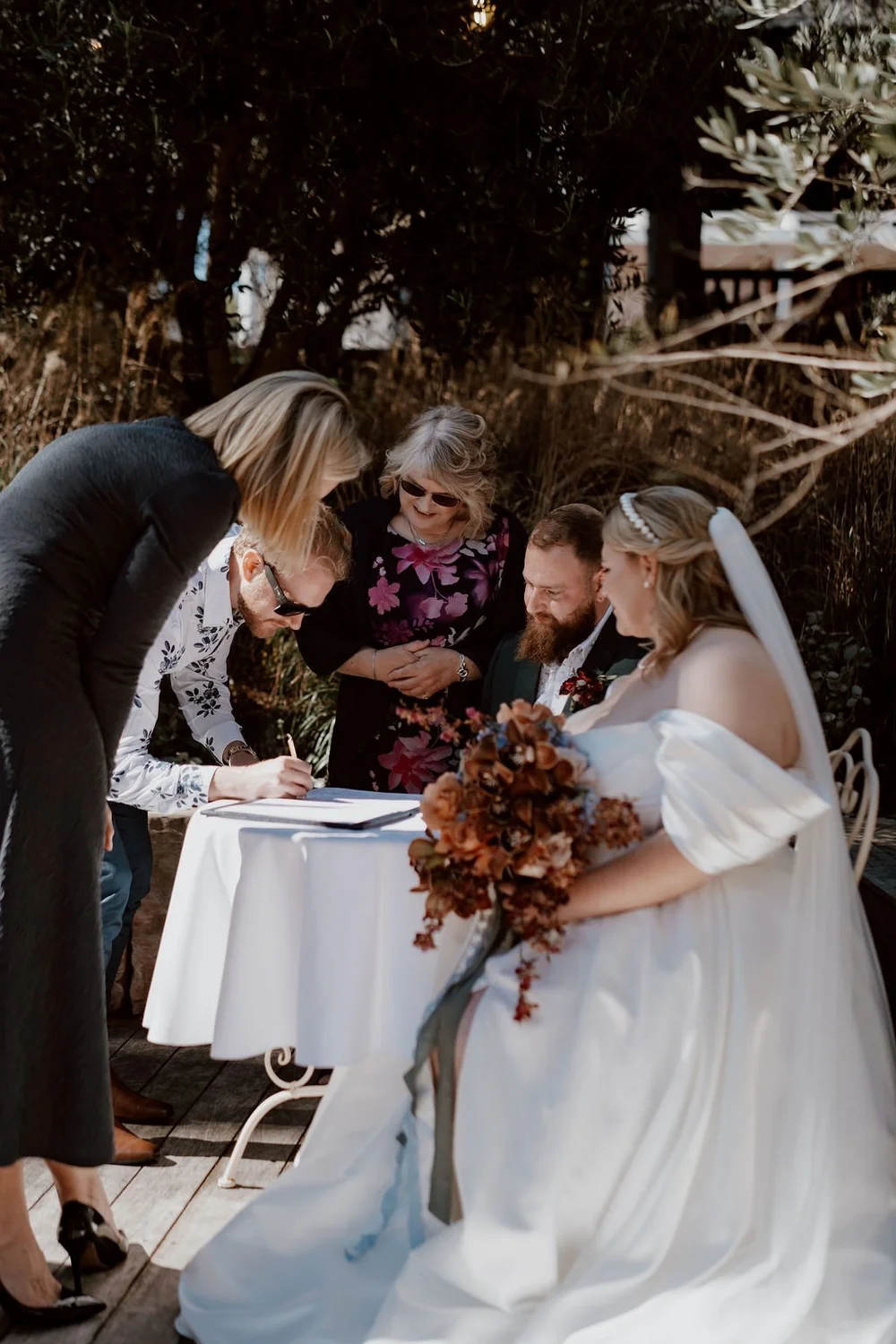 A bride in a white wedding dress holding a bouquet, seated outside, signing a document as friends and family gather around her during a wedding ceremony.