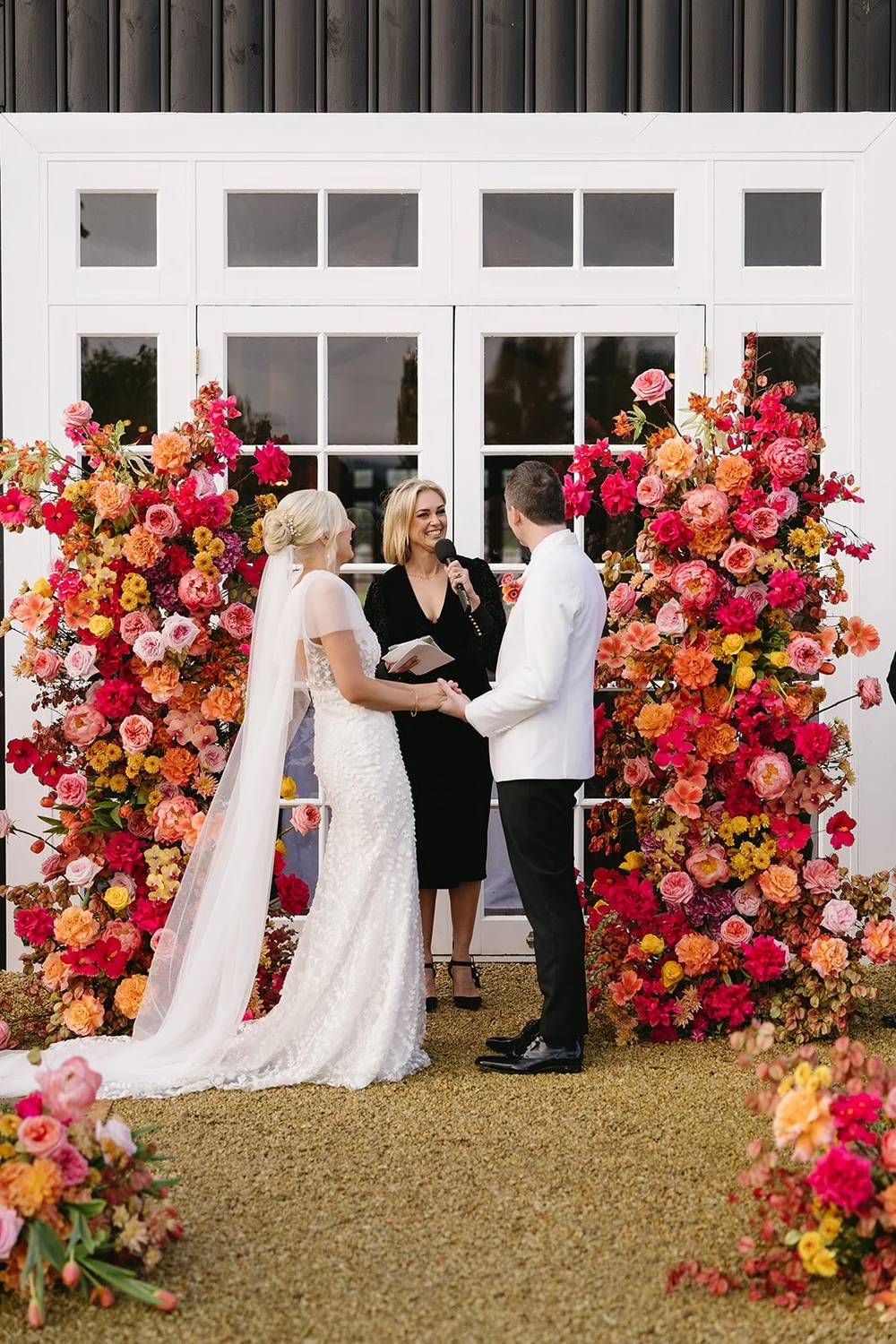 A wedding ceremony with a bride and groom holding hands before an officiant, surrounded by colorful floral arrangements, in front of a white building with glass doors.