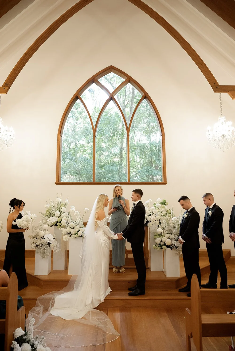 A wedding ceremony inside a church with the bride and groom holding hands, an officiant reading, and bridesmaids and groomsmen standing nearby, with large windows and floral arrangements.