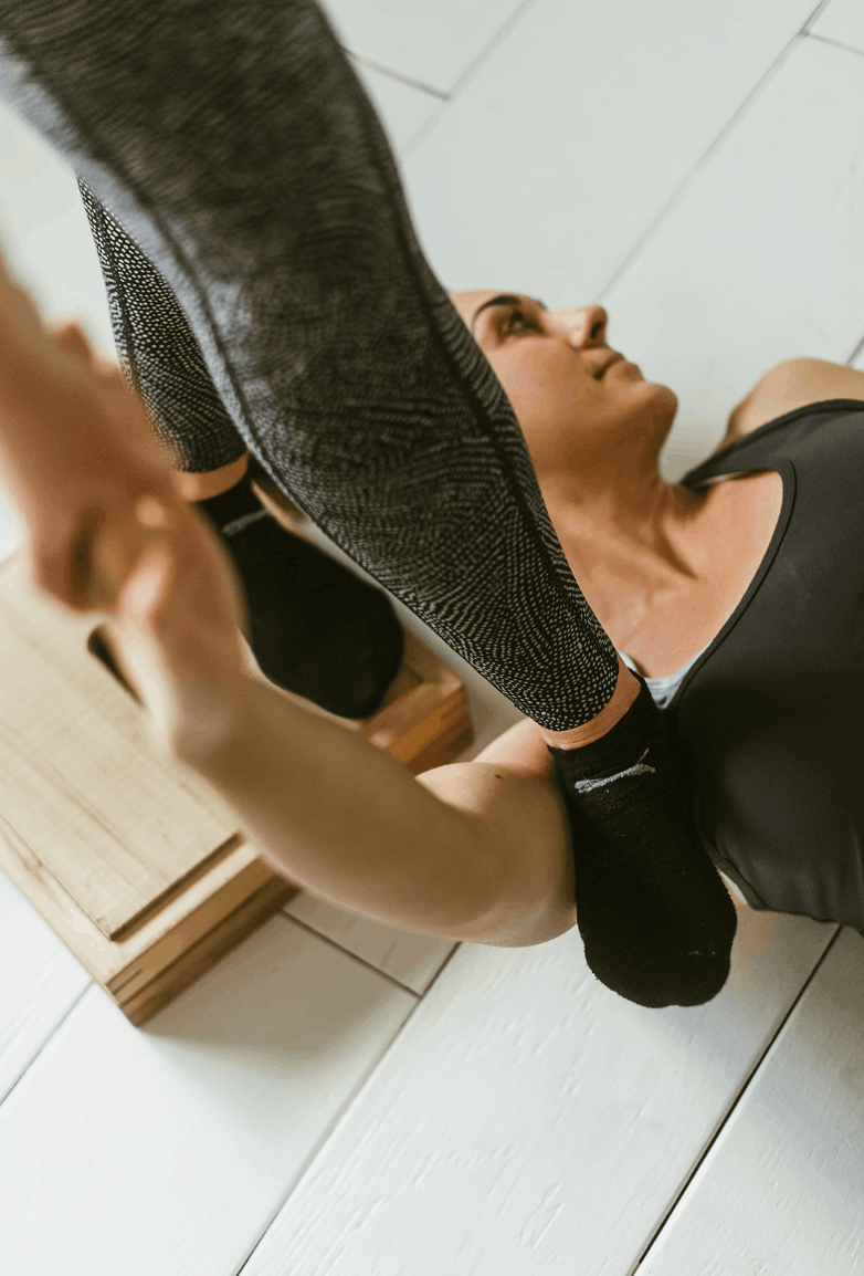A woman lying on the floor with her arms raised, holding a dumbbell, wearing a black athletic top, with a focus on her upper body and face, on a wooden floor.