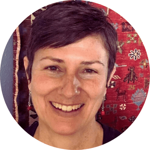 A smiling woman with short dark hair, earrings, and a nose piercing, in front of a colorful, patterned textile background.