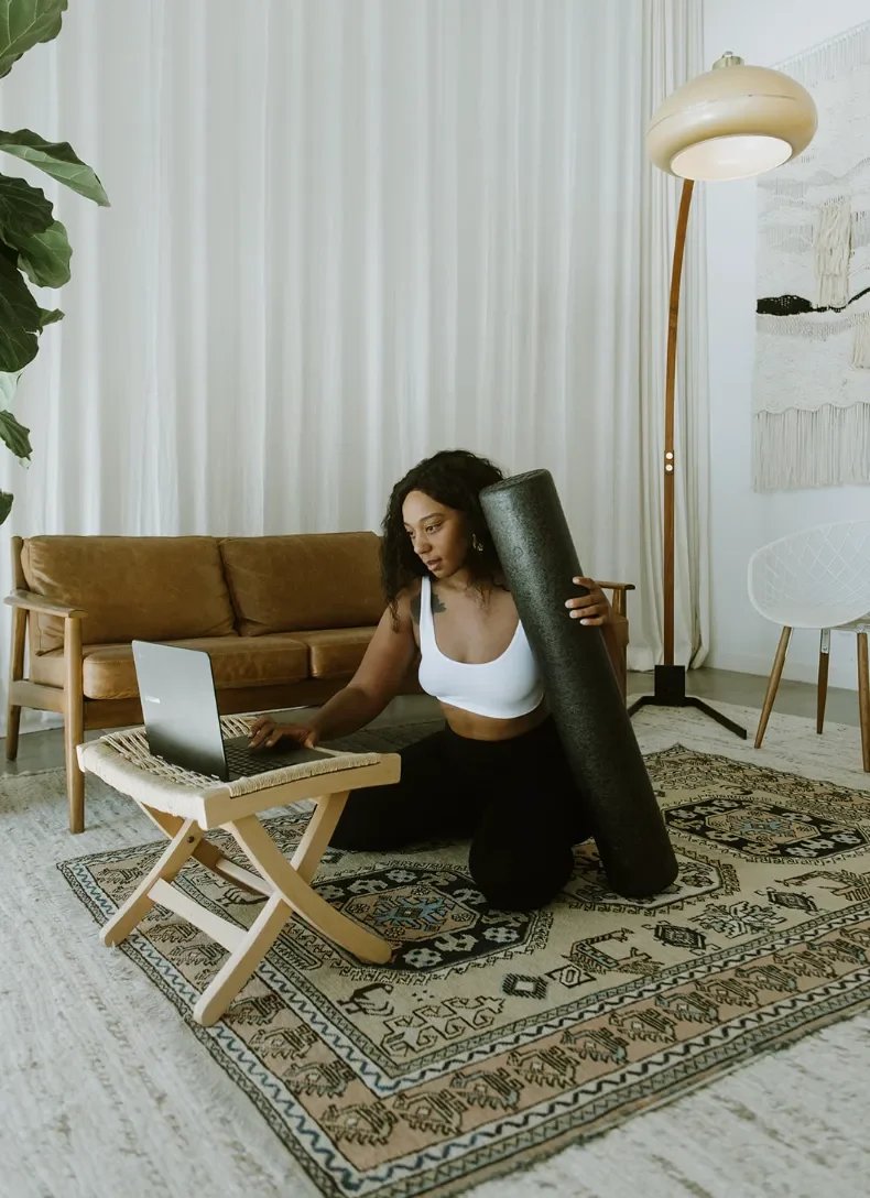 Woman on a yoga mat in a living room, looking at a laptop on a small folding table, holding a yoga mat.