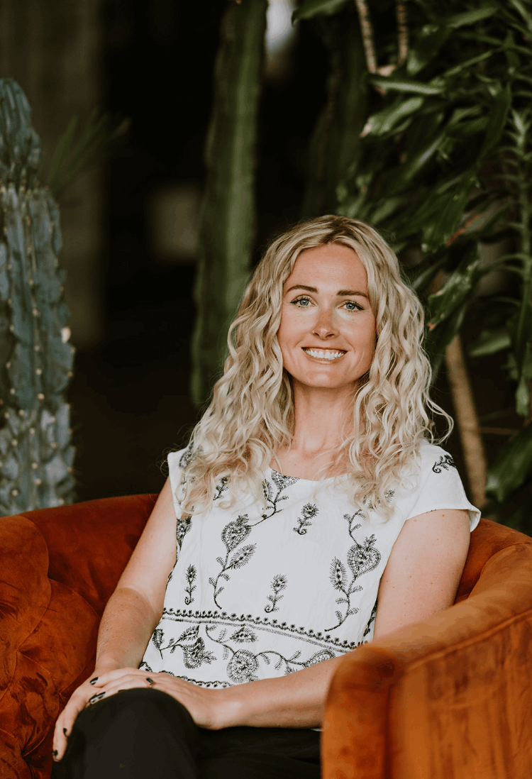 A smiling woman with curly blonde hair sitting on an orange velvet chair, surrounded by green plants.
