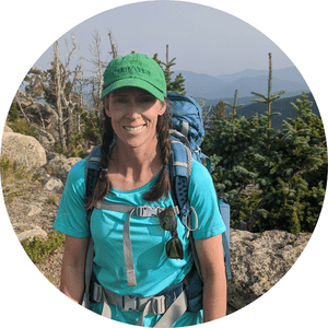Smiling woman hiking outdoors with a blue backpack, green cap, sunglasses, and outdoor clothing, mountains and trees in the background.