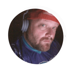 A man with a beard wearing a red cap, headphones, and a dark sports jersey or jacket, taking a selfie against a dark background.