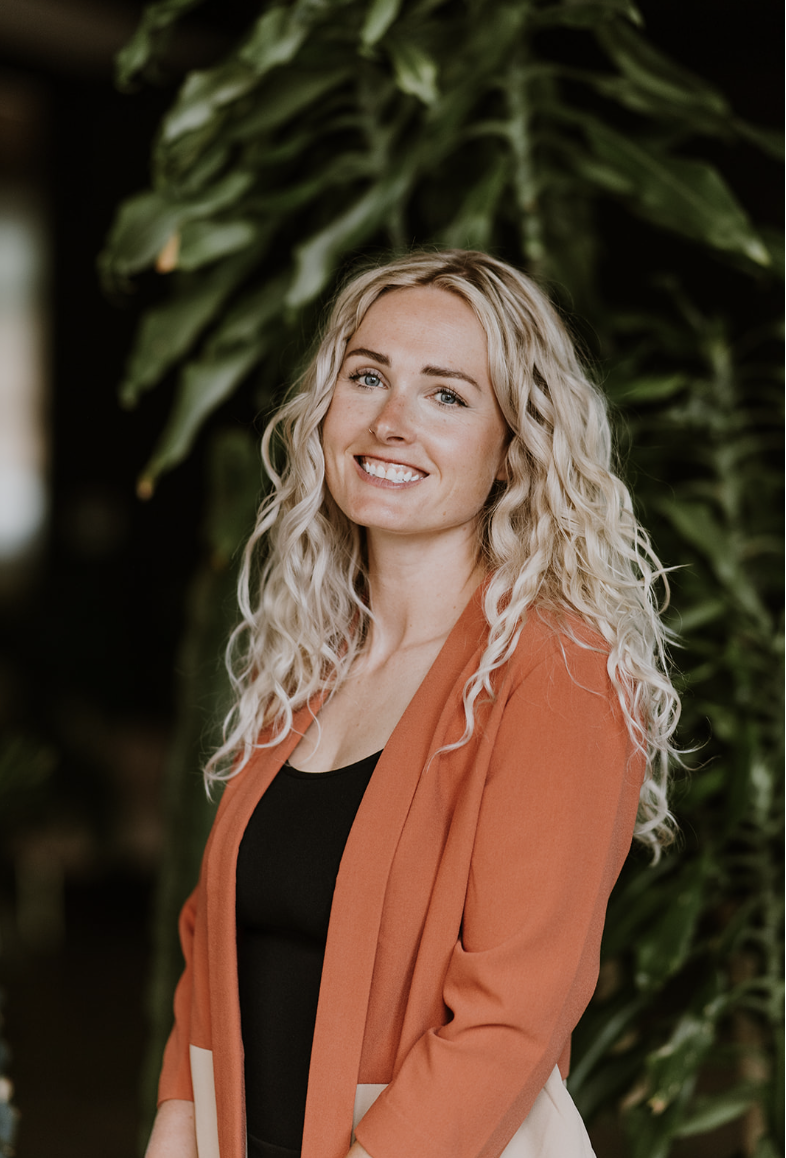A young woman with long, curly blonde hair (Julia) smiling at the camera, standing in front of large green tropical plants, wearing an orange blazer over a black top.