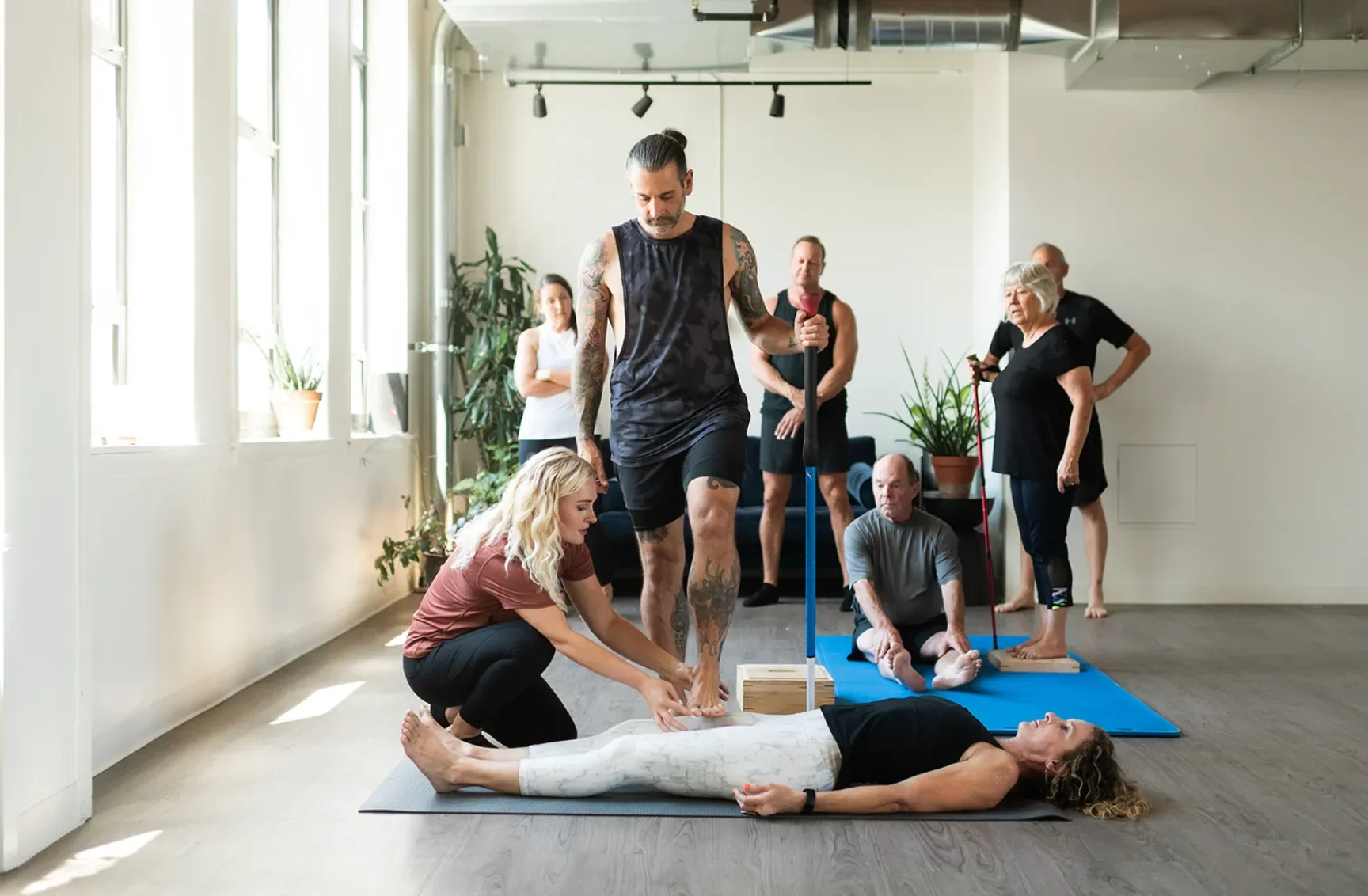 A woman is lying on a yoga mat during a group yoga or therapy session, with a woman and a man supporting her legs.