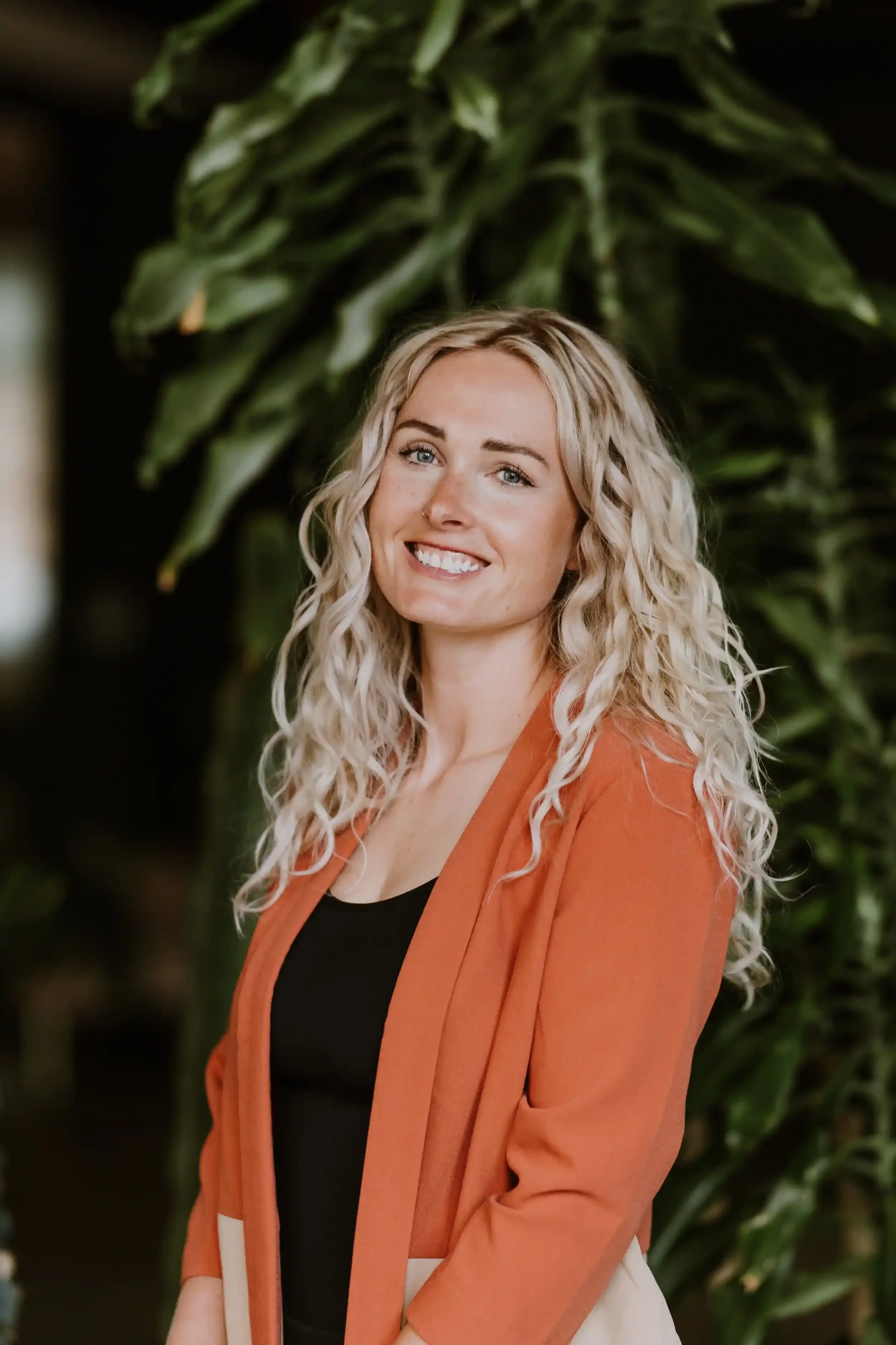 A young woman with long, curly blonde hair, smiling, wearing an orange blazer over a black top, standing in front of green foliage.
