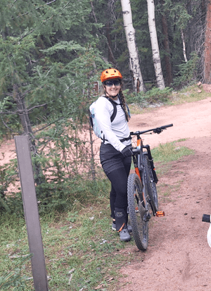 A woman in outdoor cycling gear with a helmet, sunglasses, and a backpack, standing next to a mountain bike on a dirt trail surrounded by trees.