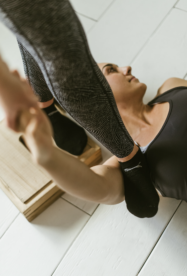 A woman lying on her back on a wooden floor, reaching up to shake hands with a person standing above her, wearing athletic clothing and socks.