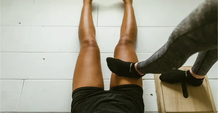 Person lying on the floor with legs extended, receiving a massage or therapy on the calf muscles from a practitioner wearing gray pants and black shoes.