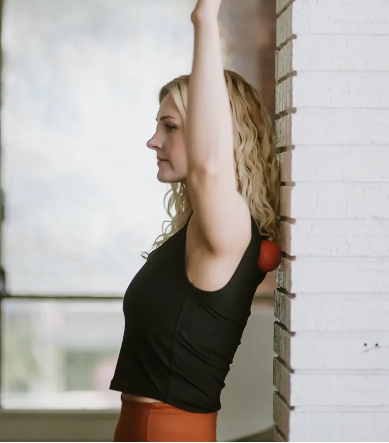 A woman with blonde curly hair leaning against a brick wall with one arm raised above her head, wearing a black sleeveless top and rust-colored pants, with a red exercise ball tucked under her arm.
