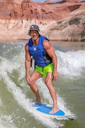 A man riding a wakeboard on a river, surrounded by red canyon cliffs, wearing a blue life vest, yellow swim shorts, a gray camouflage bucket hat, and no shoes.