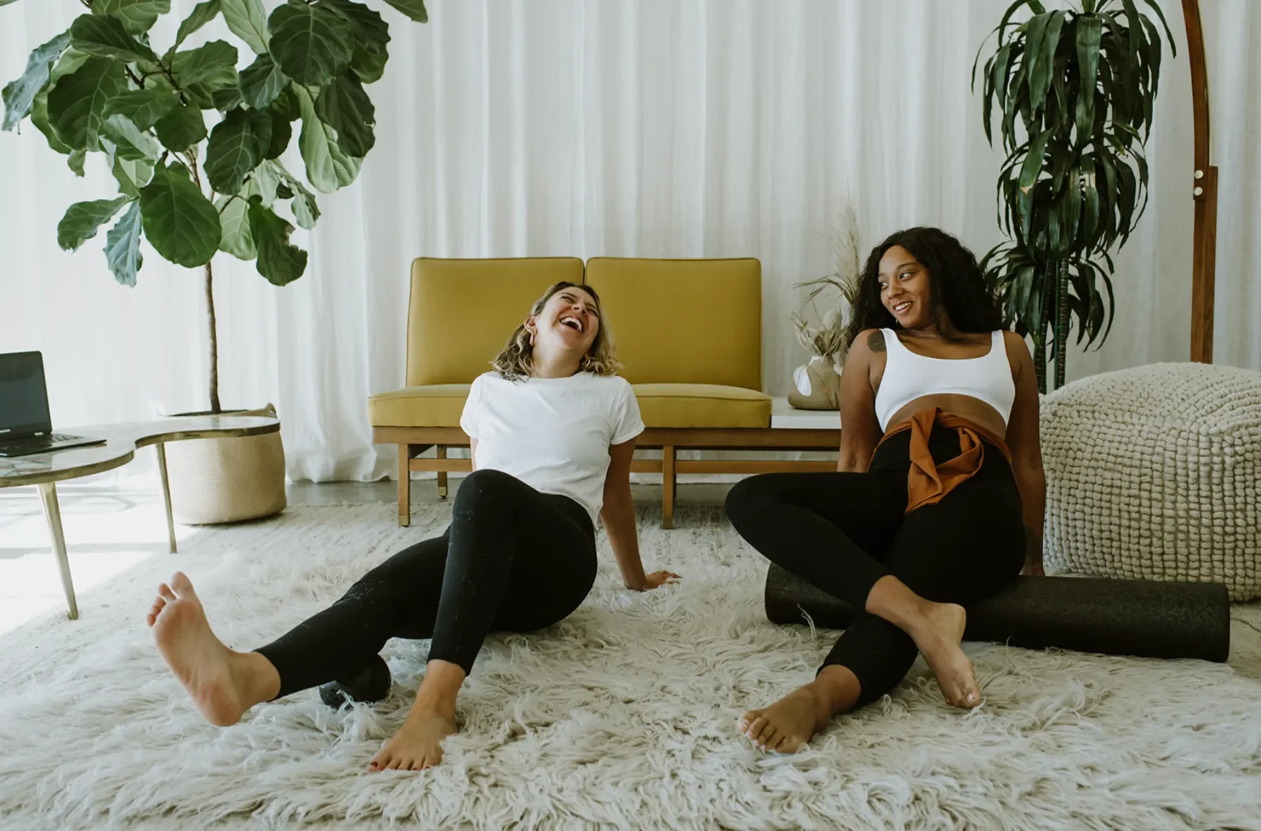 Two women sitting on a fluffy white carpet, laughing and enjoying each other's company in a bright, modern living room with large green plants and minimal decor.