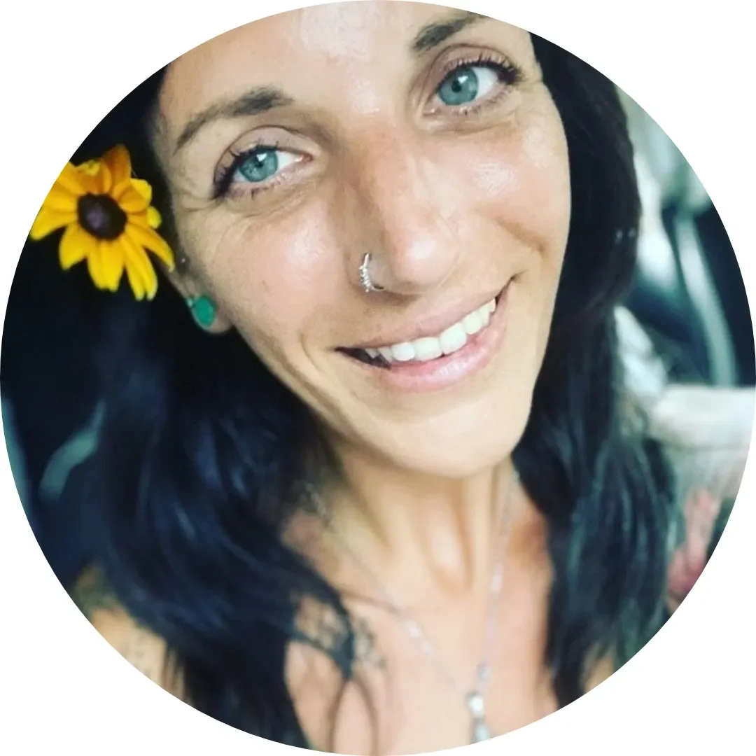 A woman with blue eyes, dark hair, a yellow flower in her hair, a nose piercing, and a black lace top smiling at the camera.