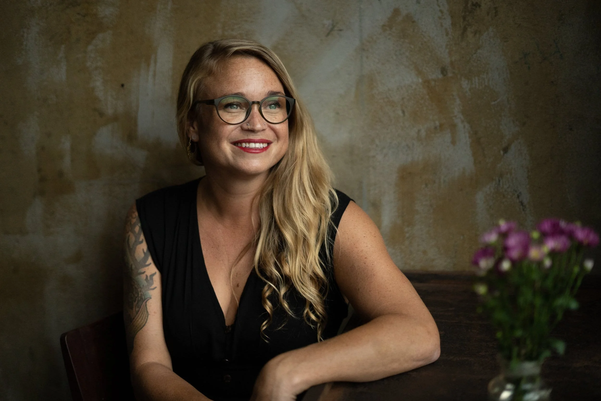 A smiling woman with long blonde hair, wearing glasses and a black sleeveless top, sitting at a wooden table with a vase of purple flowers. She has tattoos on her left arm and is looking to her left, in front of a textured beige wall.