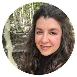 Portrait of a woman with wavy dark hair smiling outdoors in a wooded area.