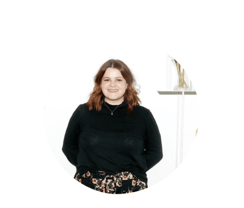 A woman with shoulder-length brown hair, wearing a black top and floral pants, standing in front of a white background with a window and glass vase in the background.