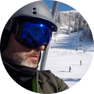 Close-up of a man in ski gear wearing a helmet and ski goggles, with a snowy ski slope and trees in the background.