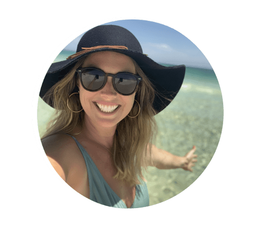 Woman smiling on the beach wearing sunglasses and a large sunhat, with clear ocean water in the background.