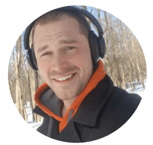 A man outdoors in a wooded area wearing headphones, an orange hoodie, and a black jacket, smiling at the camera with snow and trees in the background.