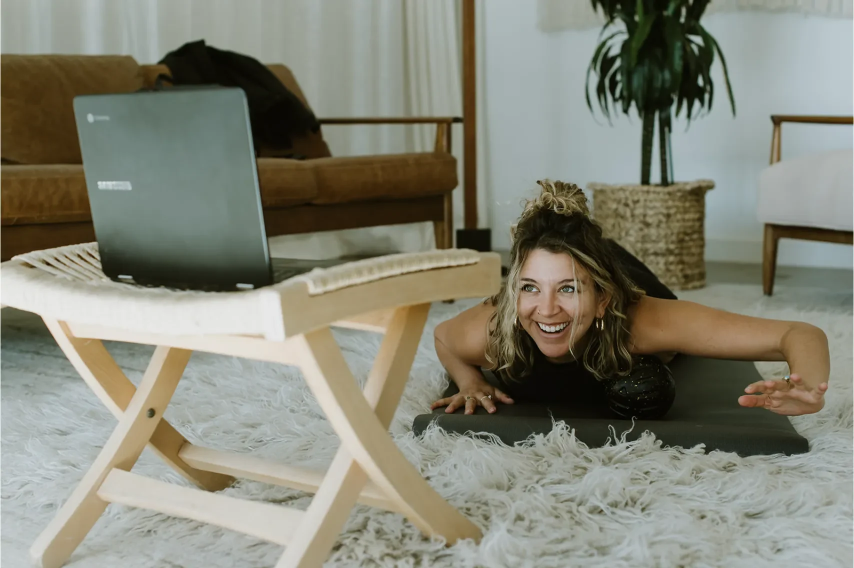 A woman doing a yoga pose on a black mat on a fluffy white rug in a cozy living room, smiling and looking at a laptop on a small wooden table.