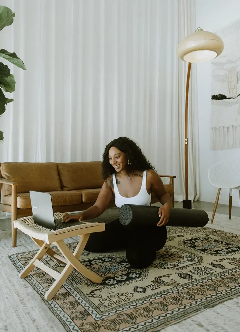 A woman in a white top and black pants is practicing yoga in a living room, using a laptop.