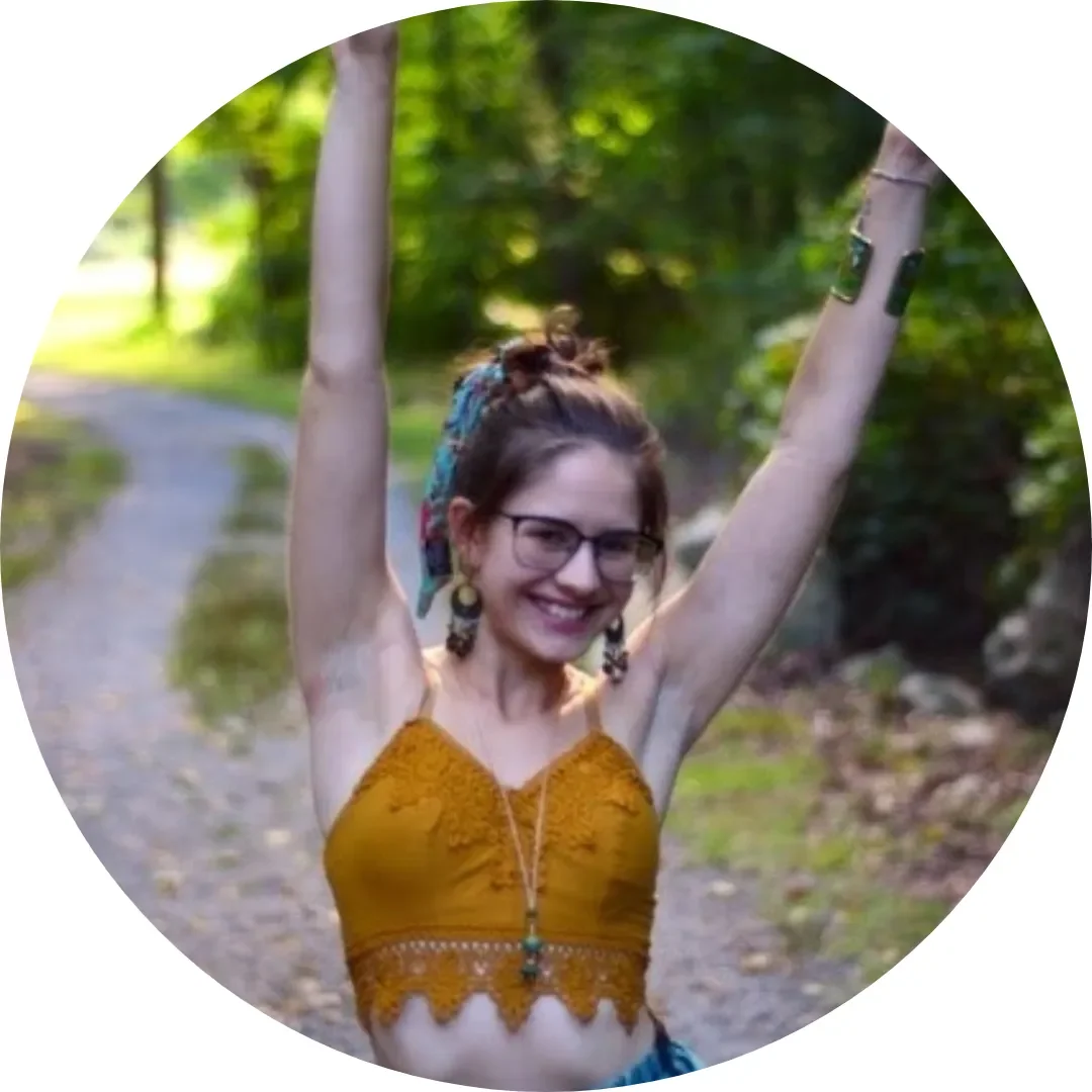A young woman smiling and raising her arms in a forested outdoor setting.