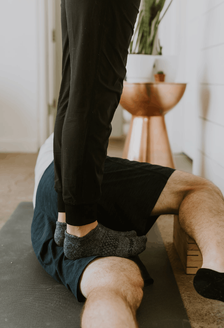 Person performing a yoga pose on a mat, with another person assisting or practicing alongside in a home setting.