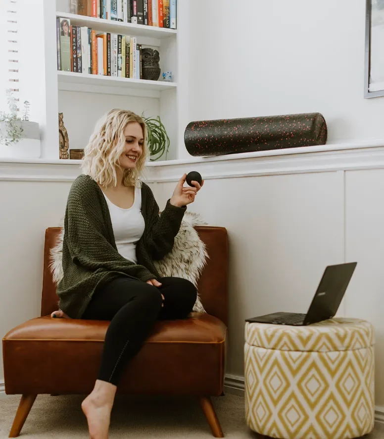A woman sitting on a brown leather chair with white fluffy pillow, smiling, holding a small black ball, with a laptop on a patterned ottoman nearby, in a room with white walls and a bookshelf in the background.