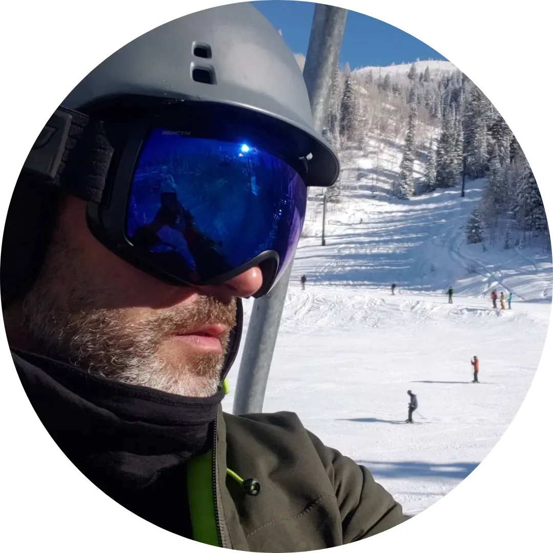 A man wearing ski goggles and a helmet taking a selfie on a snowy ski lift with a mountain and skiers in the background.