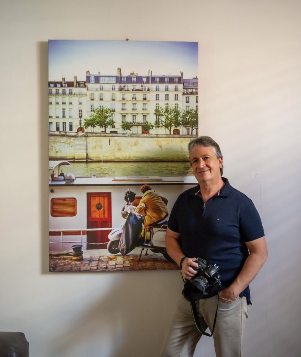 Mike Annese, founder of Blink Gallery standing indoors in front of a large Paris cityscape photography print, holding a camera, smiling, wearing a dark polo shirt and beige pants.