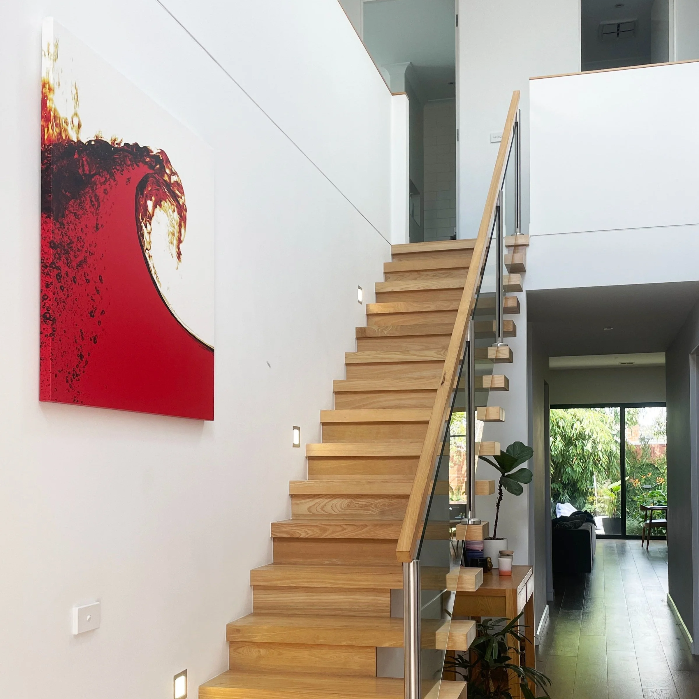 Interior view of a modern home featuring a wooden staircase with a metal and glass railing, contemporary artwork on the white wall, and a living area with large windows showing greenery outside.