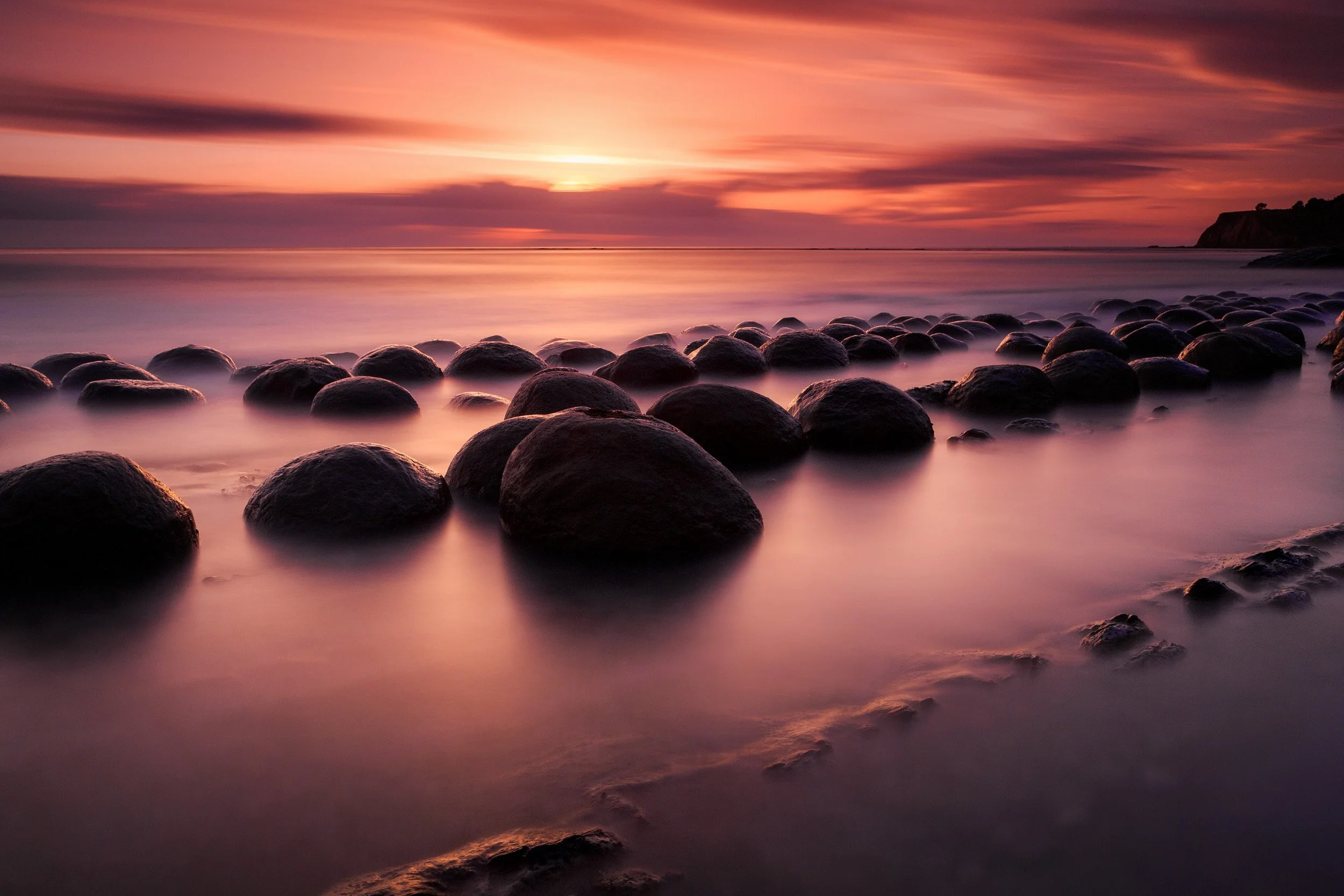 Rows of round boulders emerge from silky long-exposure surf under a deep red sunset at Bowling Ball Beach on the Mendocino coast, California.
