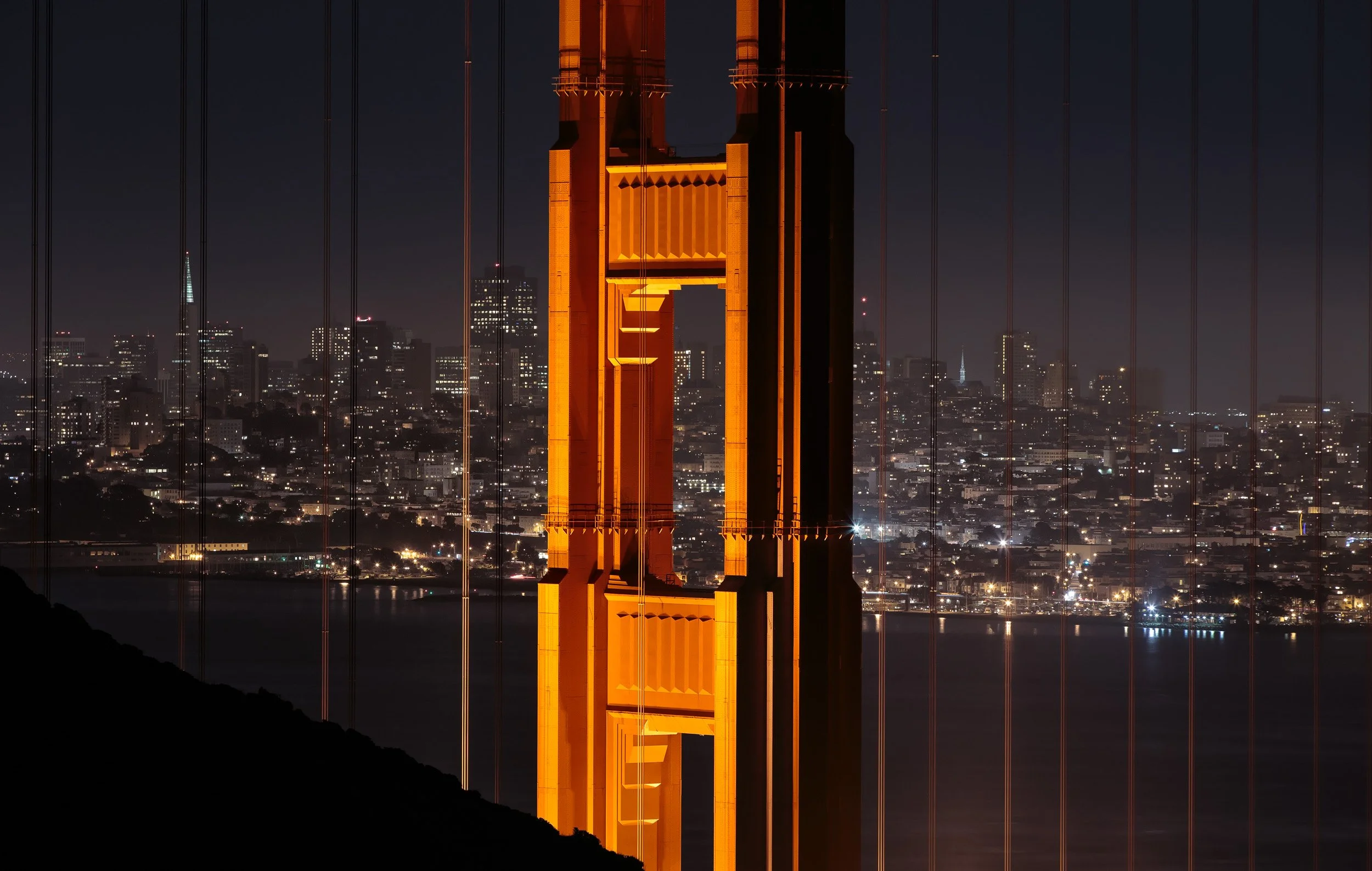 The illuminated north tower of the Golden Gate Bridge framed against the San Francisco city skyline at night.