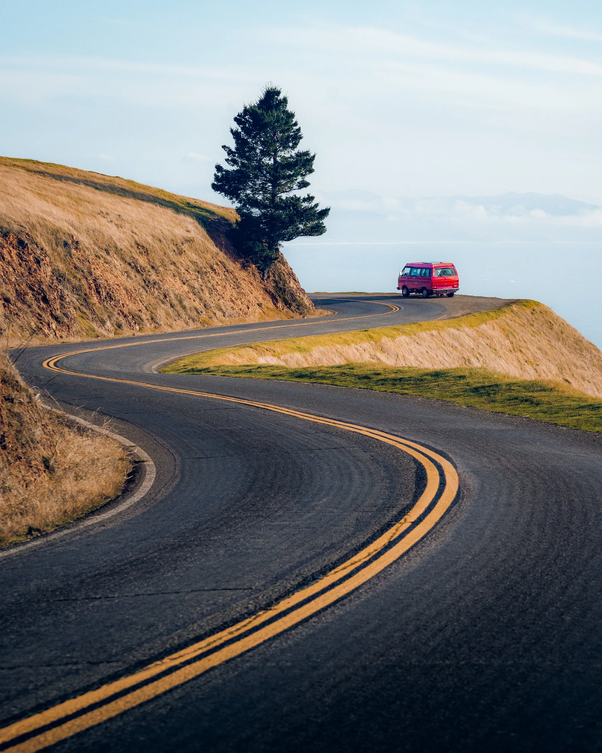A red Volkswagen van rounds a curve on a winding road above the fog on Mount Tamalpais, with the Pacific Ocean beyond.