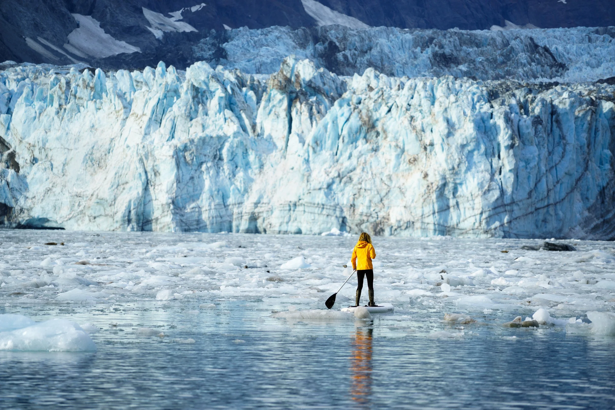 A paddleboarder in a yellow jacket stands among floating ice in front of the towering blue face of the Johns Hopkins Glacier, Alaska.