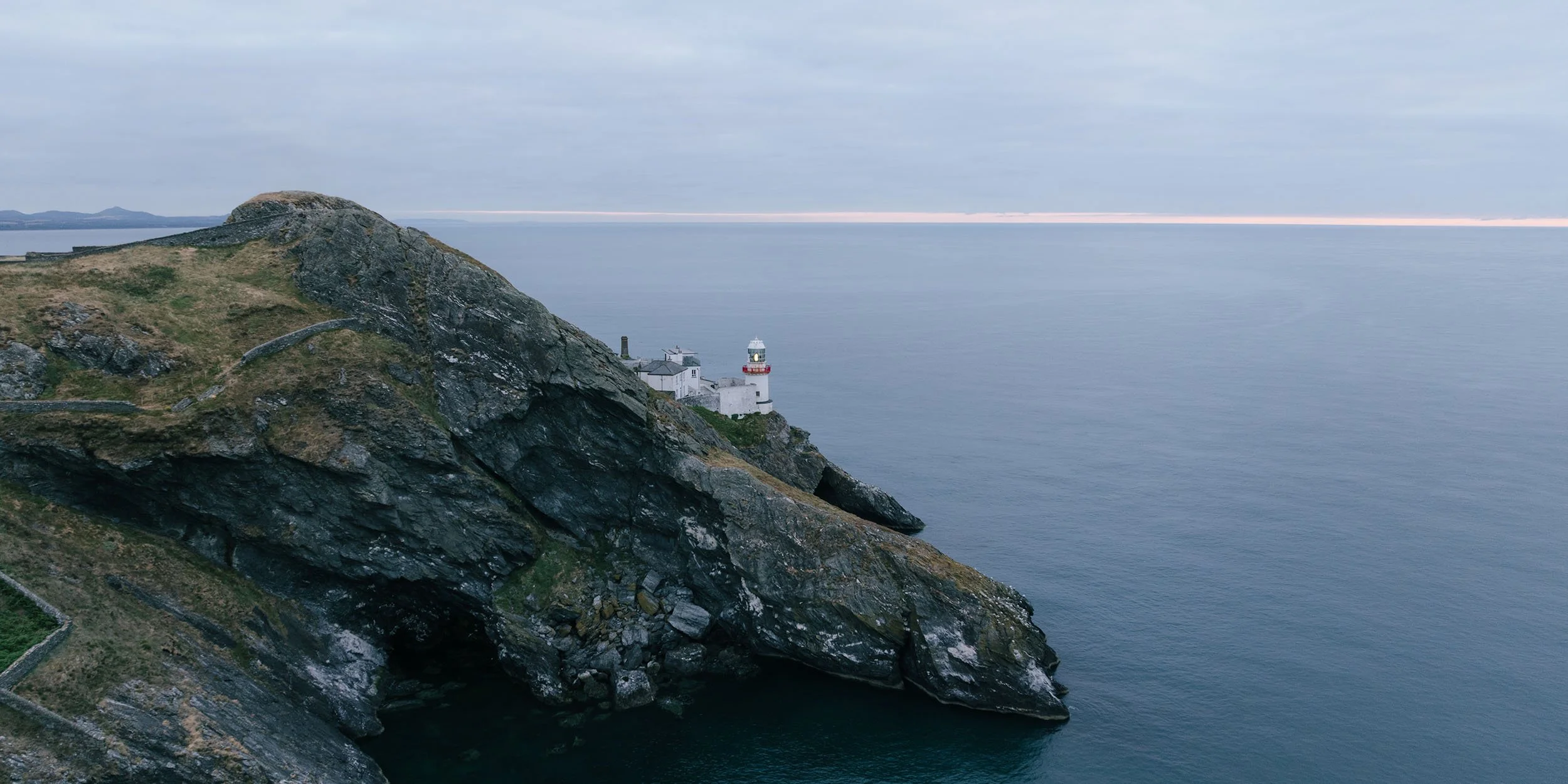 Aerial view of Wicklow Head lighthouse on a rocky headland above calm sea at dusk, County Wicklow, Ireland.