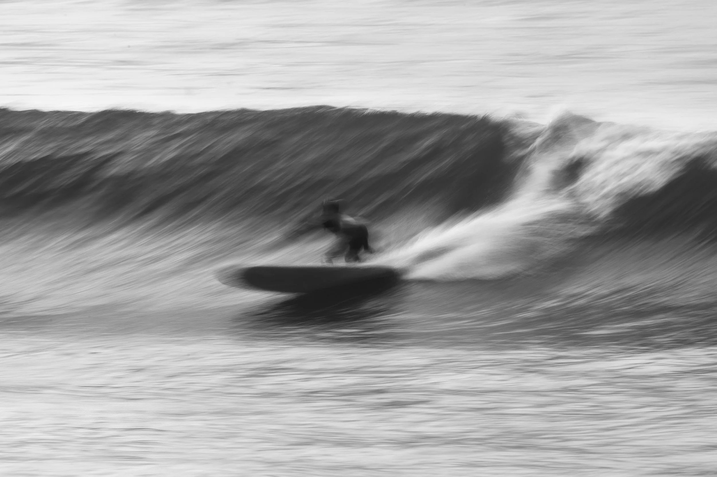 A surfer drops into a wave in a black and white long exposure, the panning motion blurring the water into streaks.