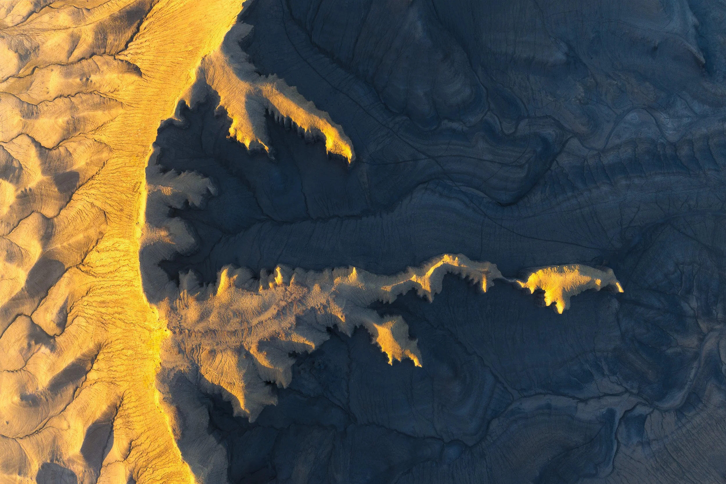 Aerial view of sunrise gold raking across grey badland canyons in an abstract pattern, Utah.